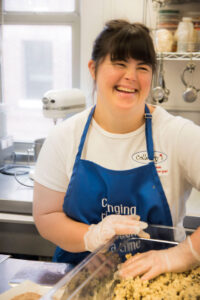 Photo shows a smiling young adult woman wearing a blue apron and food safe gloves in front of cookies on a sheet