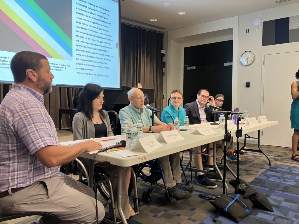 The presenters sit at the tables with the screen showing the Disability Pride Flag behind them. From left to right: Gary is seated on the side of the table, wearing a purple and white plaid shirt. Nyree is wearing a black shirt and grey cardigan. Robert, wearing a teal shirt, is speaking into the mic. Nathan is wearing a light blue shirt and looking towards the camera. Jack is wearing a black jacket and touching his glasses. Barbara is wearing sunglasses and facing forward