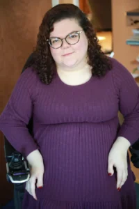 Photo of Emily Ladau, a young woman with dark hair, wearing glasses and a purple dress. 