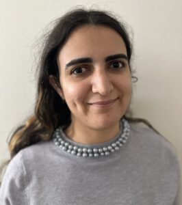 A headshot of a smiling woman with long dark hair, wearing a grey sweater with a beaded pearl collar, posed against a neutral off-white background