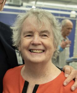 Portrait of Jini, smiling with short grey hair and wearing a vibrant orange top, in an indoor setting