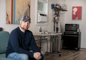 A medium shot photo showing a smiling man with a beard, wearing a navy henley shirt, jeans, and a baseball cap, sitting on a teal couch inside a brightly lit art studio.