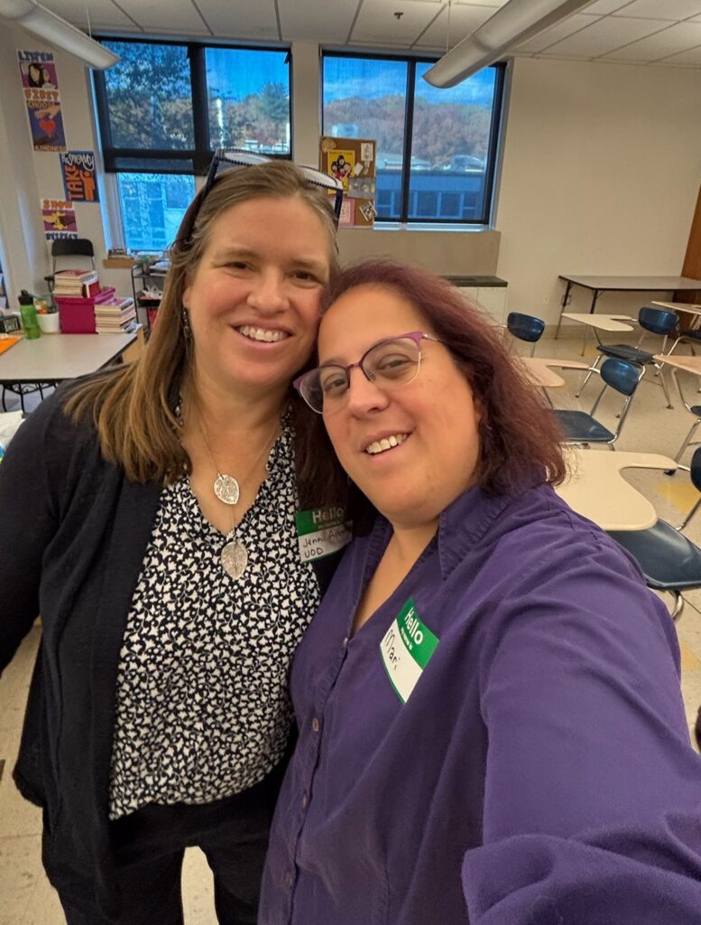 Jenni and Mari smiling together for a selfie in a classroom setting."` **Detailed Option:** `alt="Two smiling women, Jenni and Mari, taking a selfie inside a classroom with desks, windows, and posters in the background. Mari wears purple-framed glasses and a purple button-down shirt, while Jenni wears a patterned shirt, dark cardigan, and a silver leaf pendant necklace.